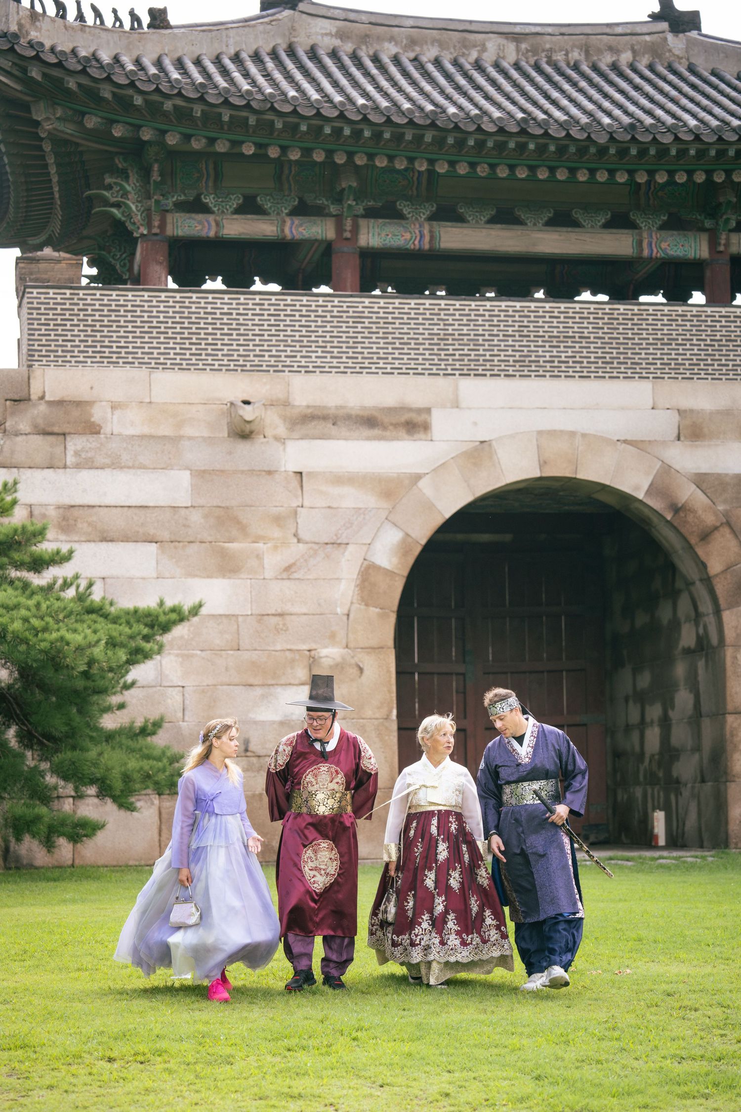 A group in traditional Korean hanbok clothing sits on the grass near an ancient palace wall with traditional tiled roofing.