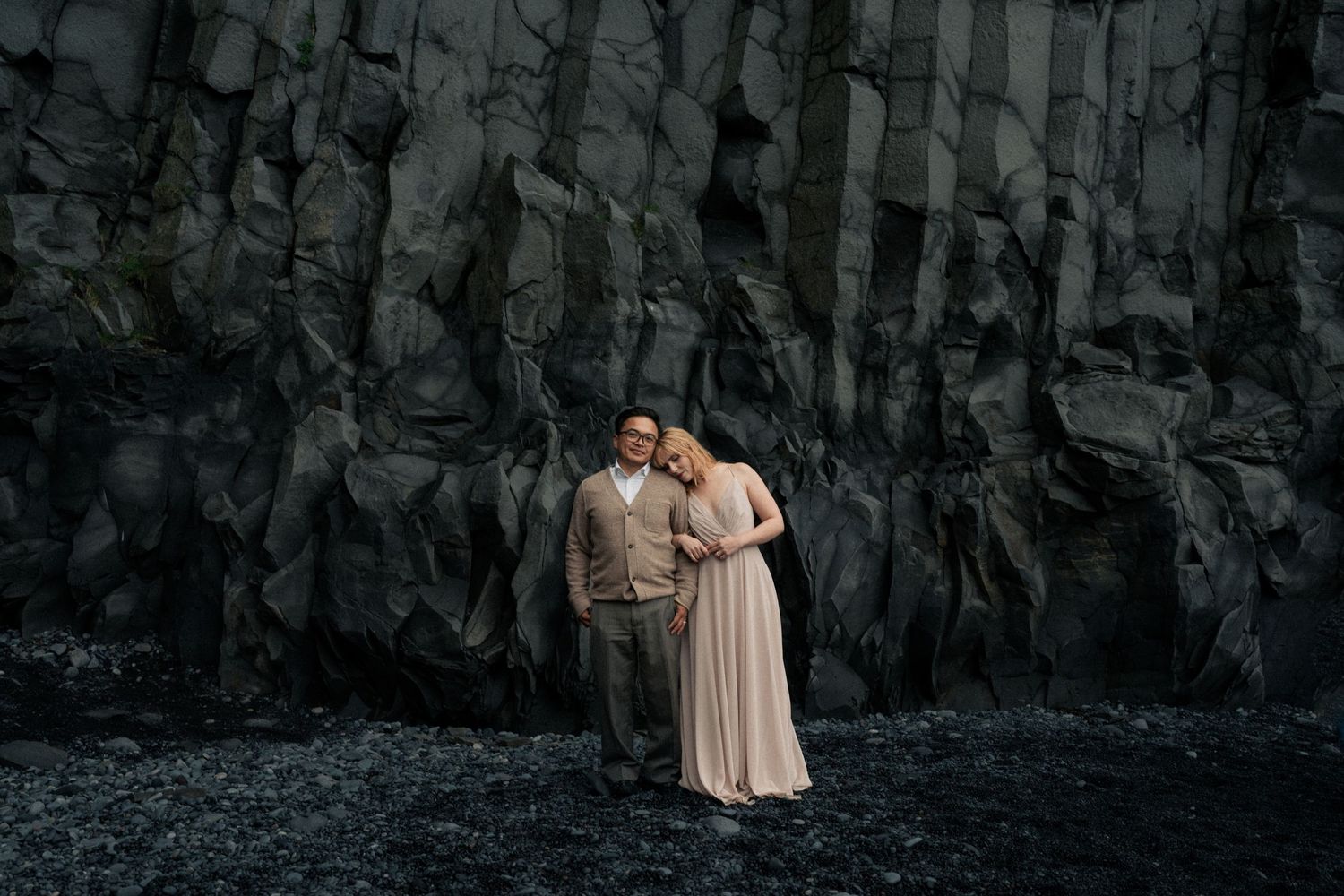 A couple embraces against dramatic dark basalt rock columns on a black sand beach.
