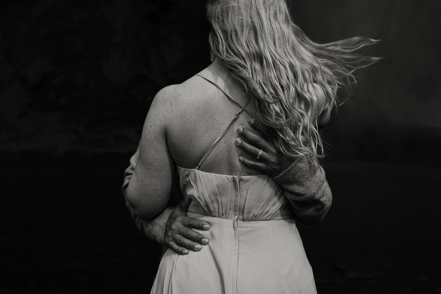 Black and white photo of a couple embracing from behind, womans blonde hair blowing in the wind.