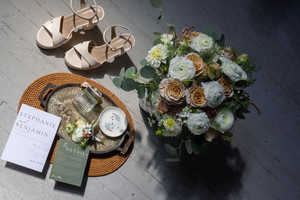 Wedding flat lay with bridal bouquet, sandals, invitation suite, and accessories on dark floor.