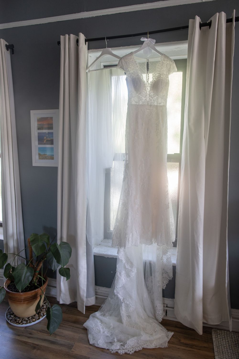 Elegant lace wedding gown hanging in window with sheer curtains and natural backlight.