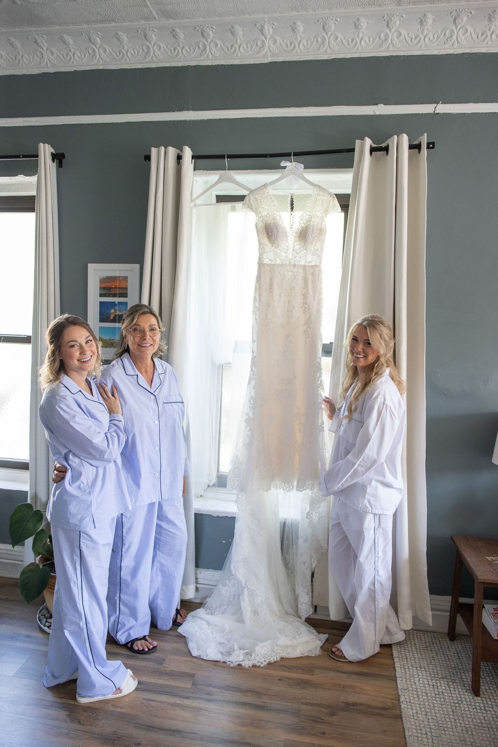 Bride and two bridesmaids in matching pajamas posing with wedding dress hanging in window.