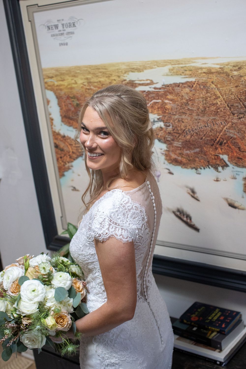 Smiling bride in lace wedding gown holding bouquet in front of vintage New York City map artwork.