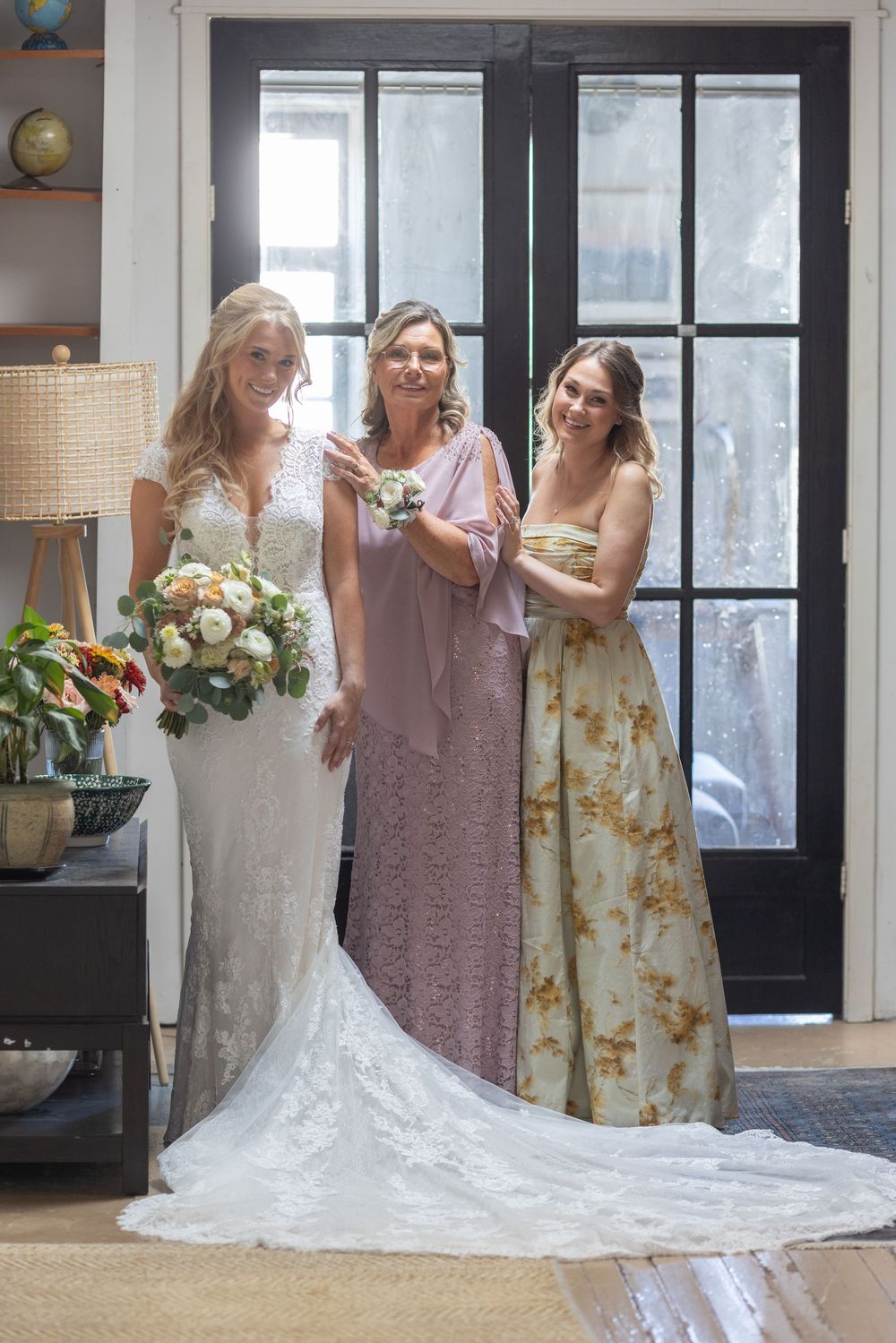 Bride in lace wedding gown poses with two women in elegant dresses near a dark French door.