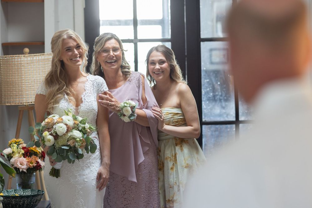 Bride in lace gown holds bouquet while smiling with two women in formal dresses near a window.