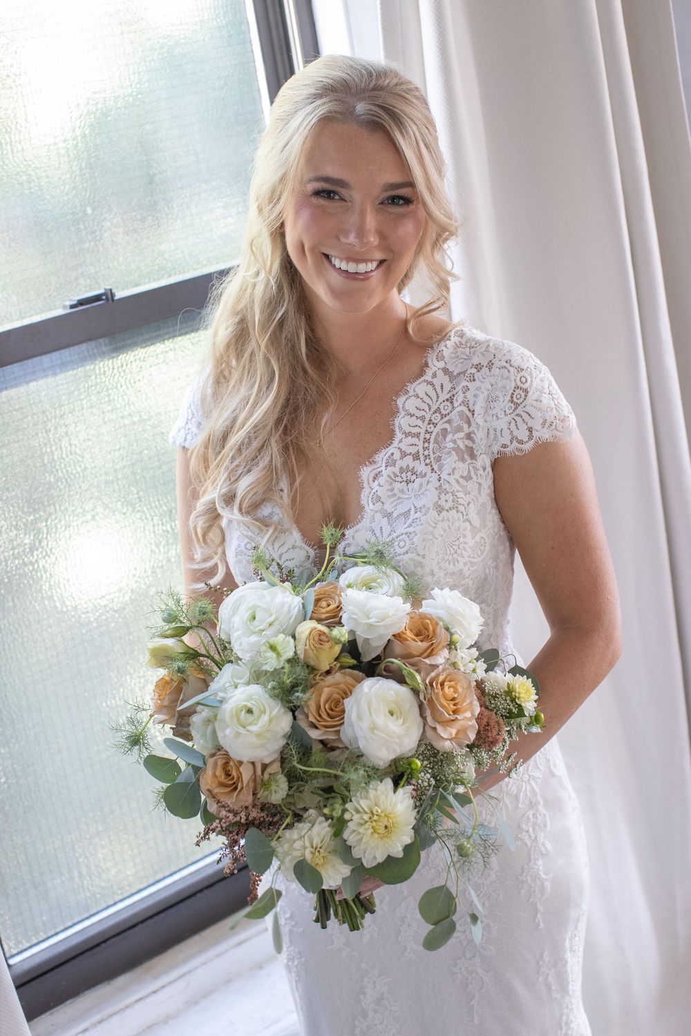 Smiling blonde bride in lace cap-sleeve gown holds peach and white floral bouquet by a window.