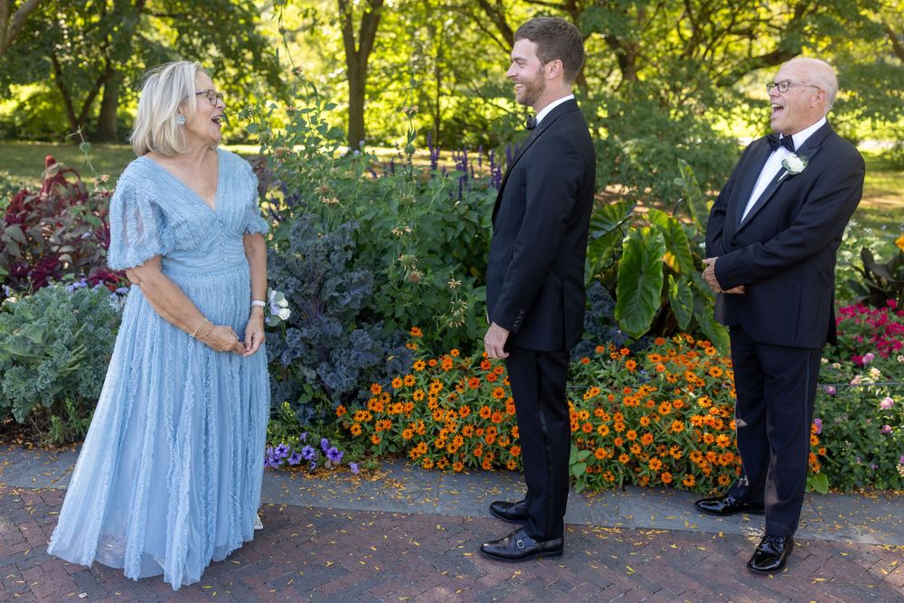 Woman in blue gown speaks with groom in tuxedo and older man in suit in a garden setting.