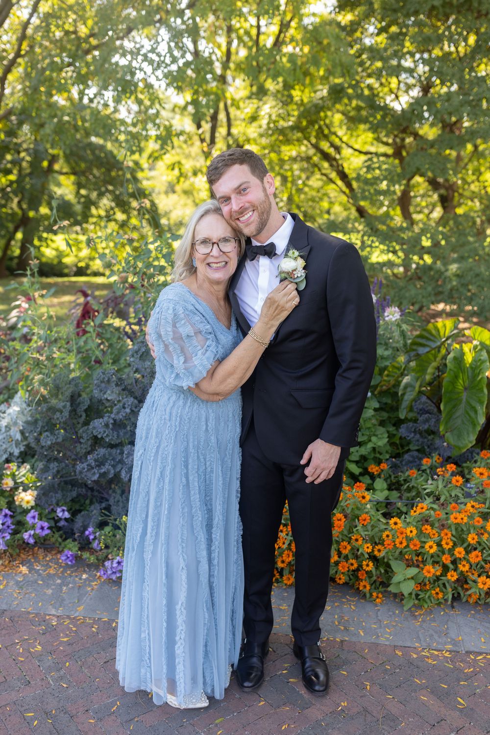 Older woman in blue lace gown embraces smiling groom in black tuxedo in a colorful garden.