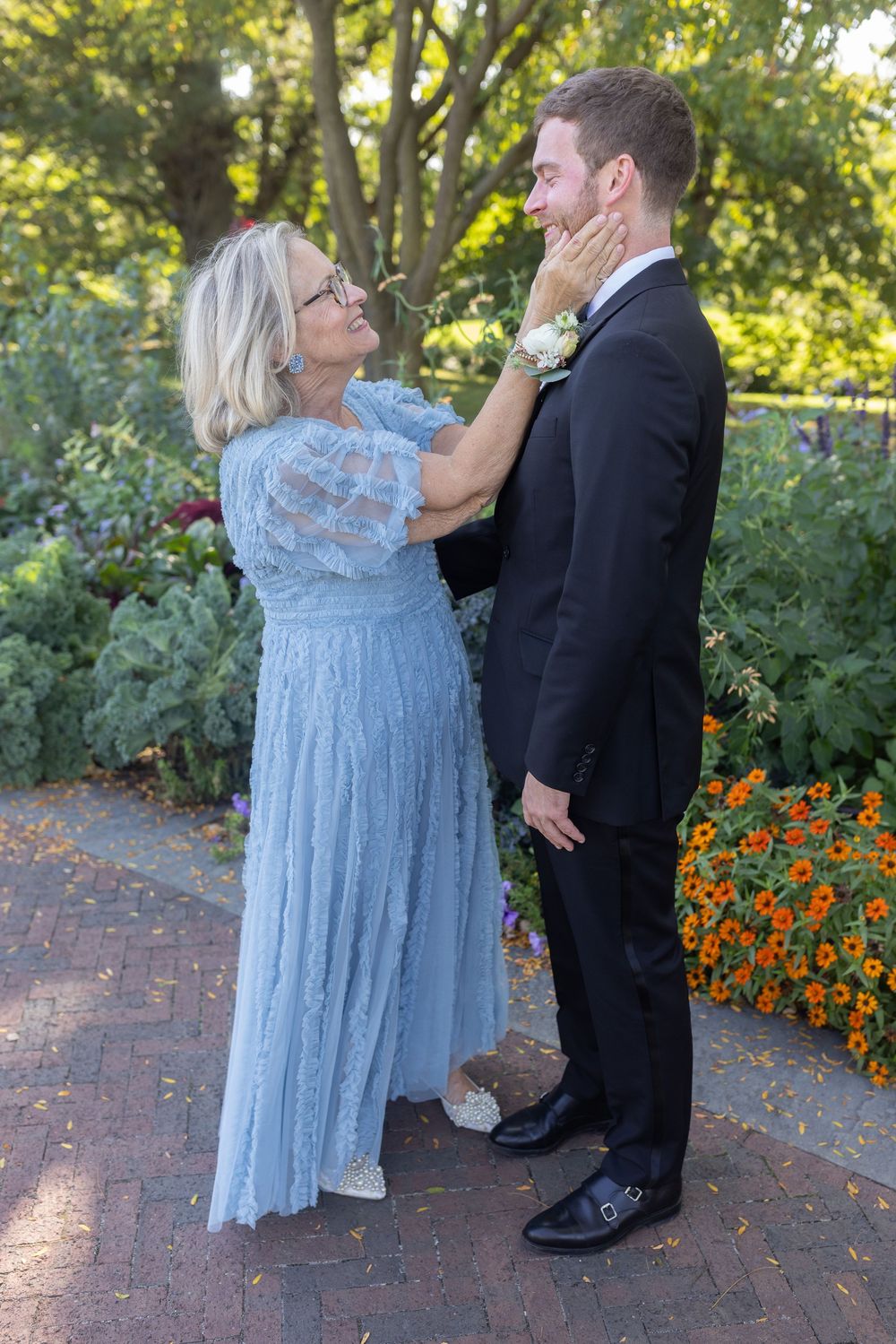 Mother in blue lace dress adjusts groom's boutonniere while standing in a vibrant garden.