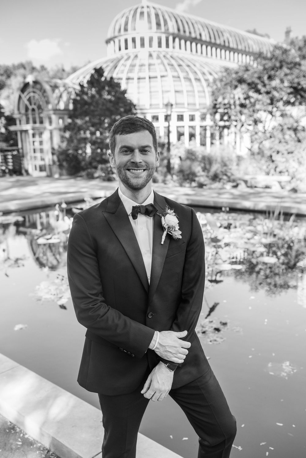 Black and white portrait of smiling groom in tuxedo standing before a glass greenhouse and pond.