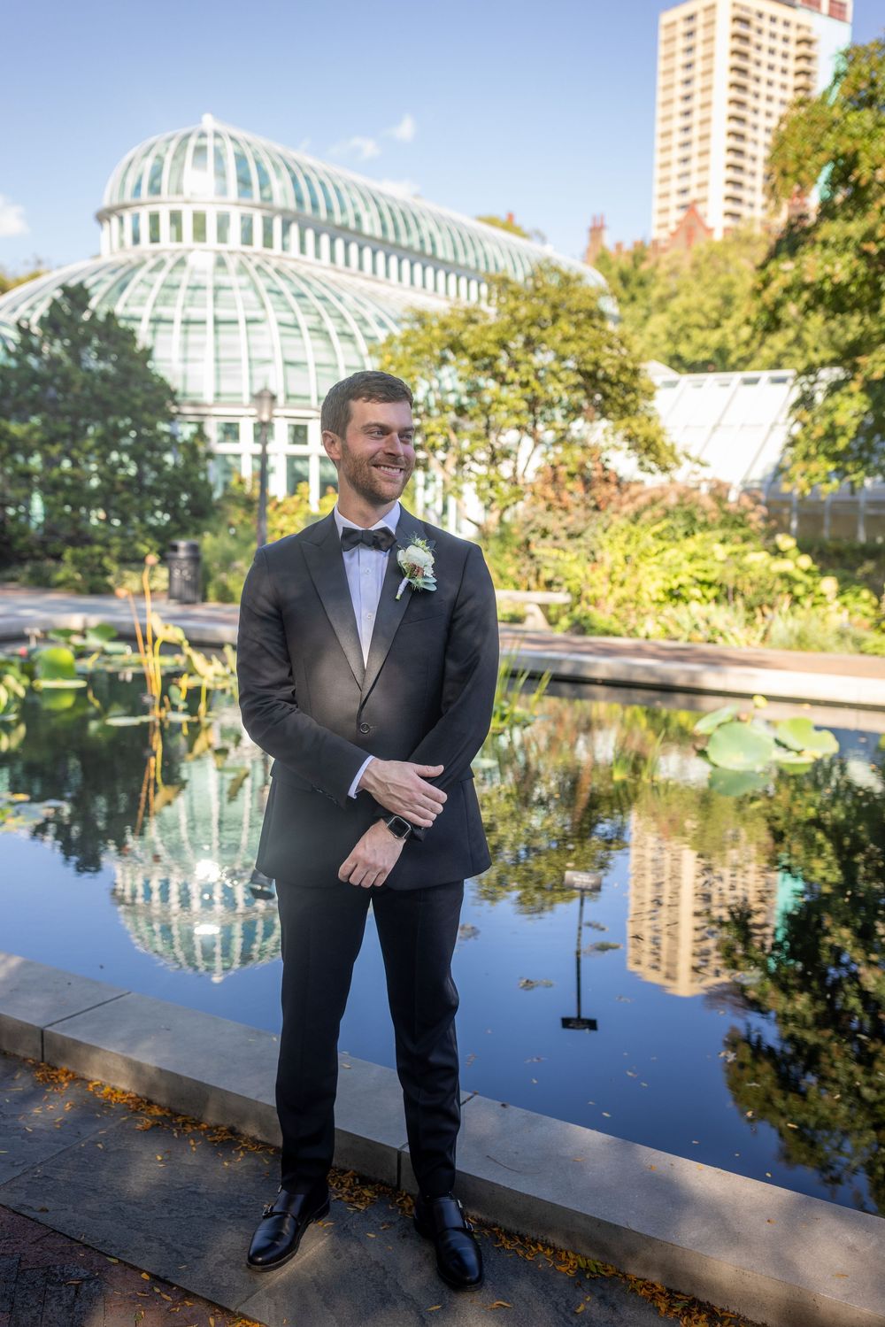 Groom in black tuxedo smiles beside a reflective pond with a domed greenhouse in the background.