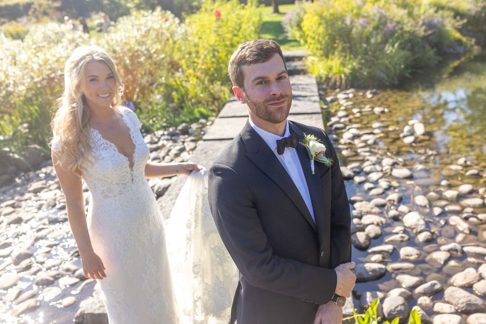 Bride in lace gown and groom in black tuxedo pose on a stone path by a rocky stream on their wedding day.