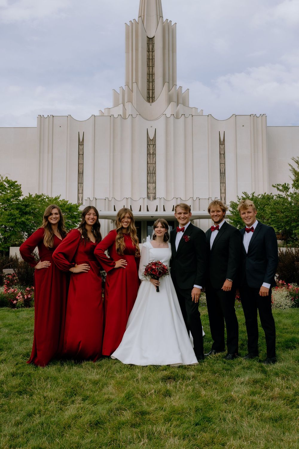 Wedding party in burgundy dresses and black suits poses in front of white temple building.