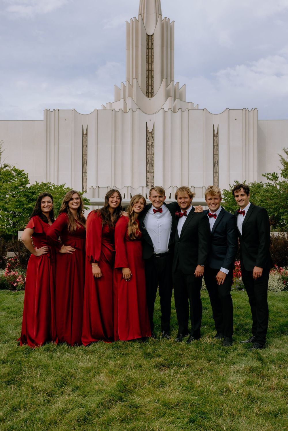 Wedding party in red dresses and black suits posing in front of a white temple building.