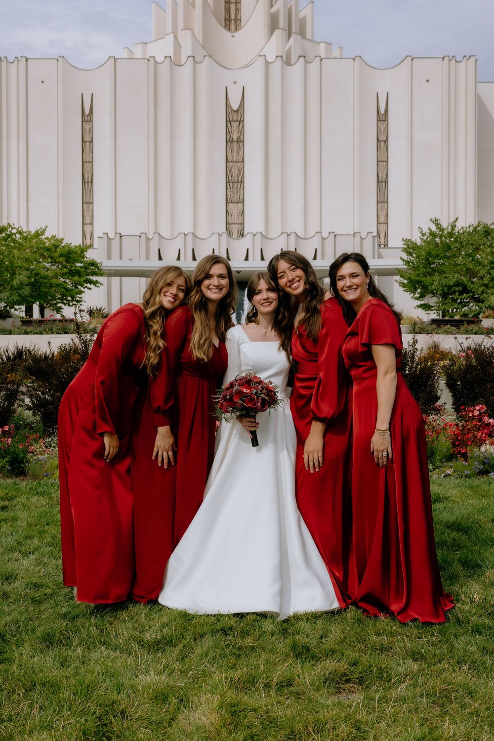Wedding party poses in front of white temple with bridesmaids wearing coordinated deep red dresses.
