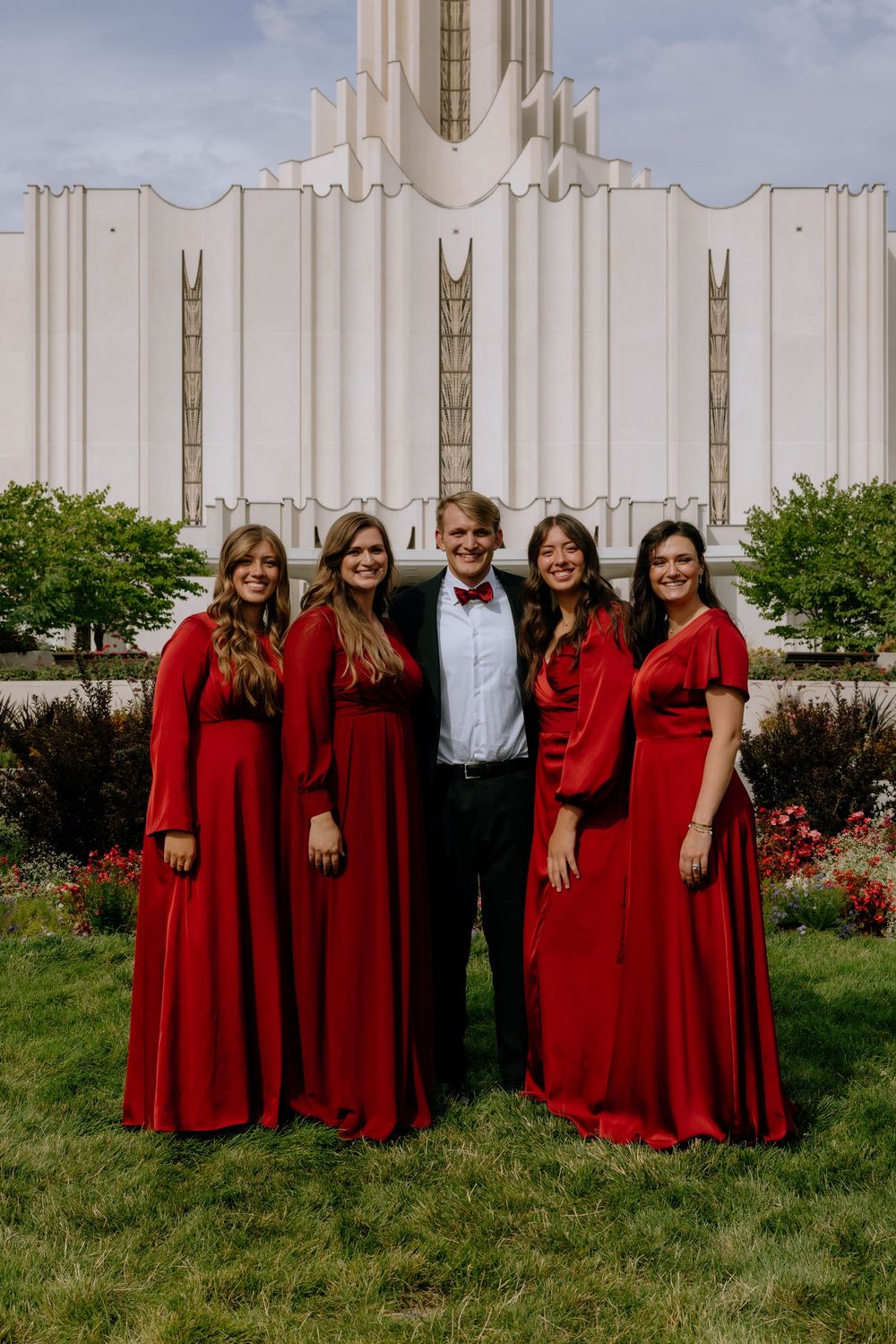 Wedding party in red bridesmaid dresses and black tuxedo pose in front of white temple building.