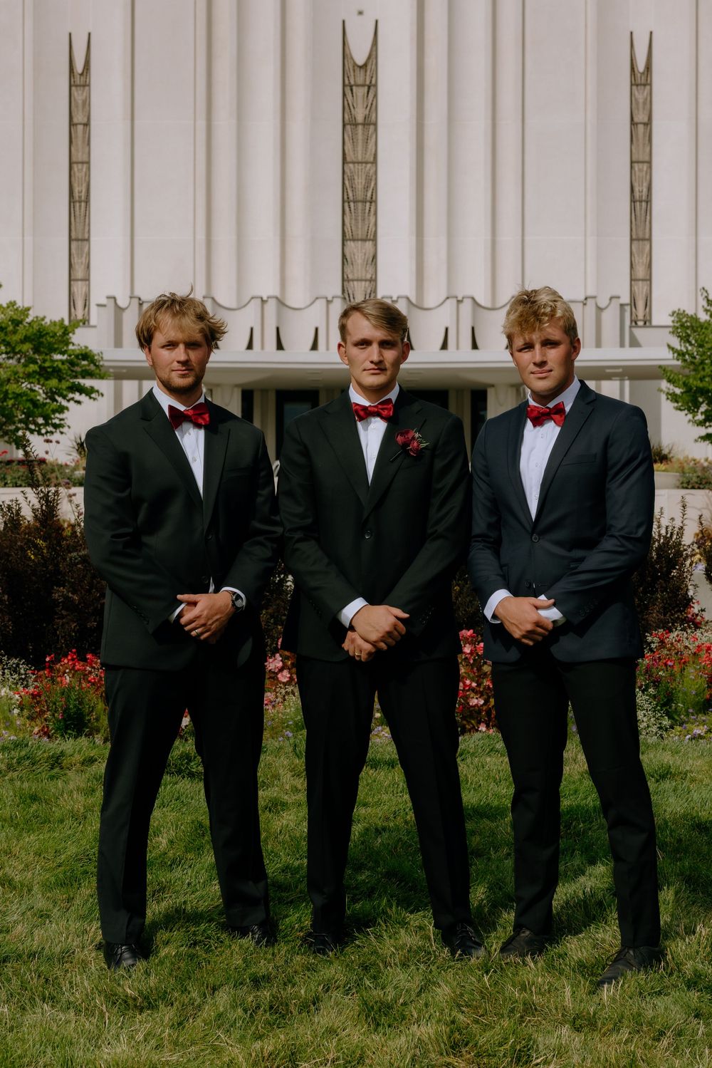 Groomsmen in black suits and red bowties pose together on grass in front of a white building.