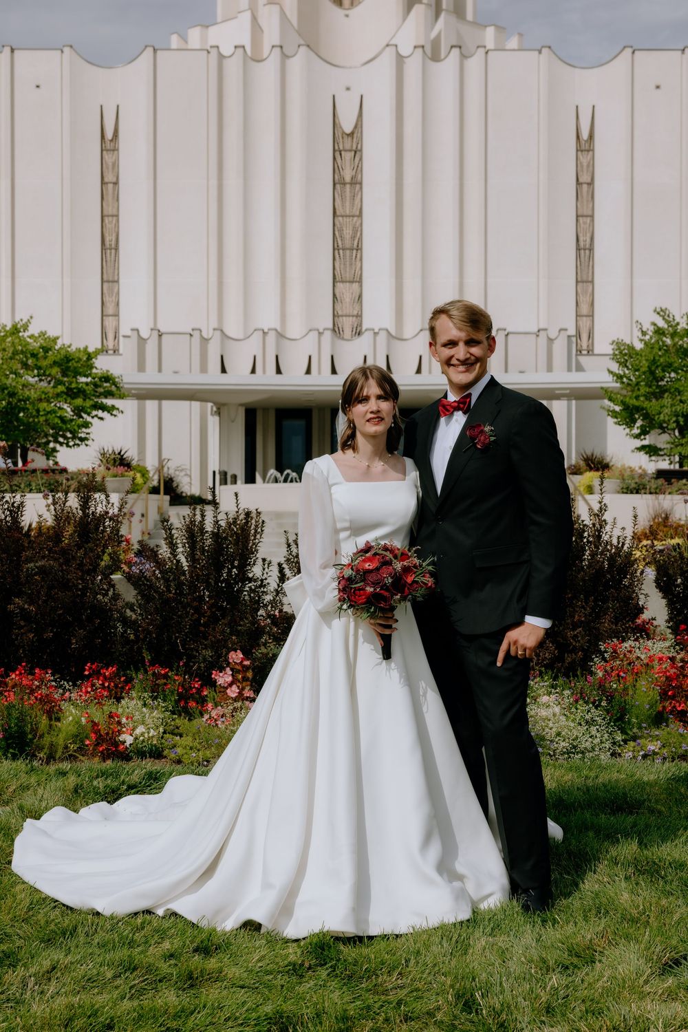 Wedding couple poses in formal attire against white temple architecture with landscaped grounds.