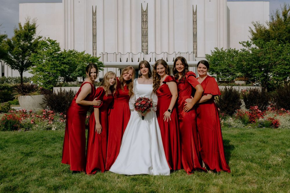 A wedding party wearing vibrant red bridesmaid dresses poses together on a grassy lawn near a white building.