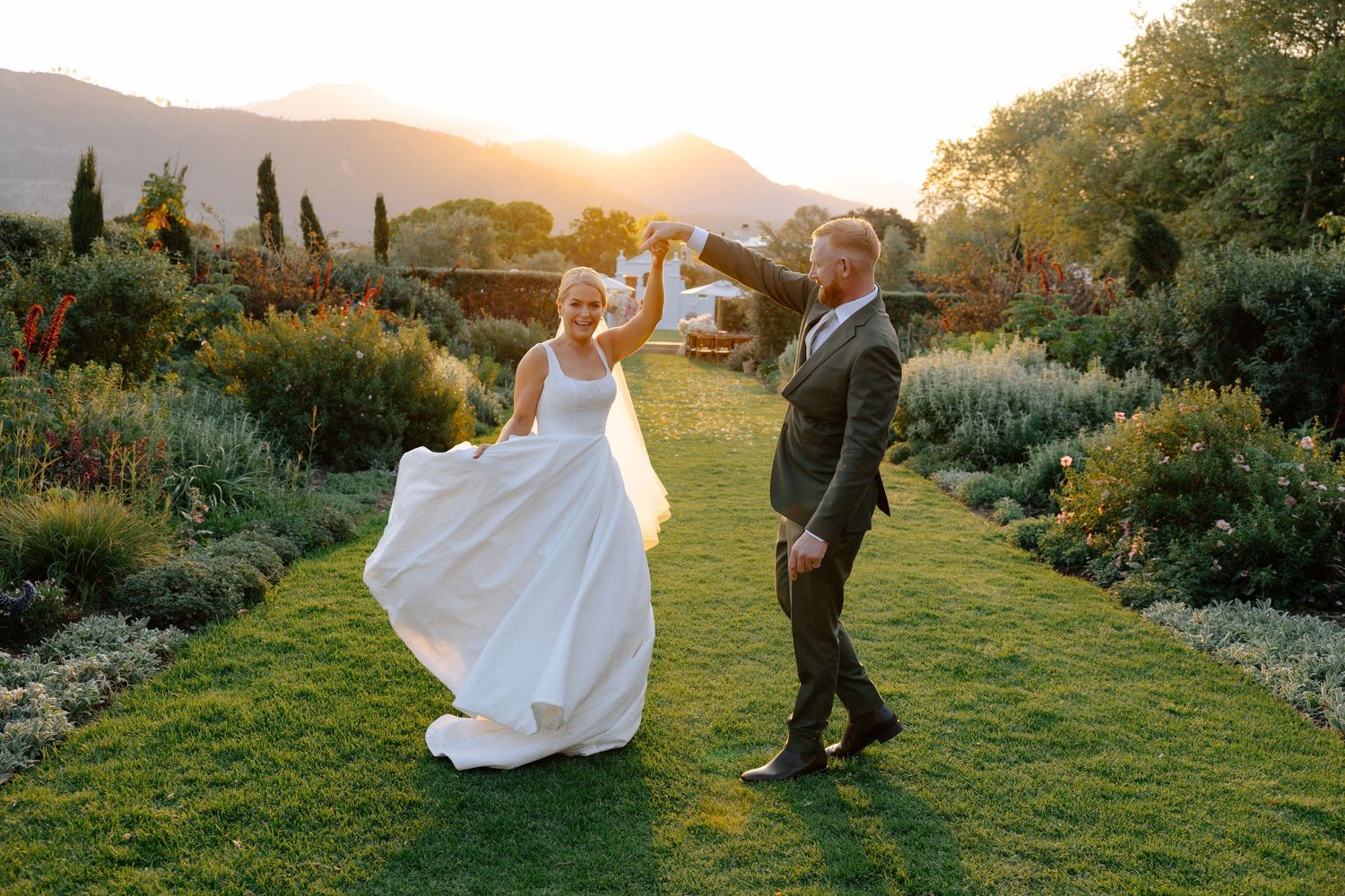 A couple dances together on a lush garden lawn during golden hour with mountains and sunset in the background.