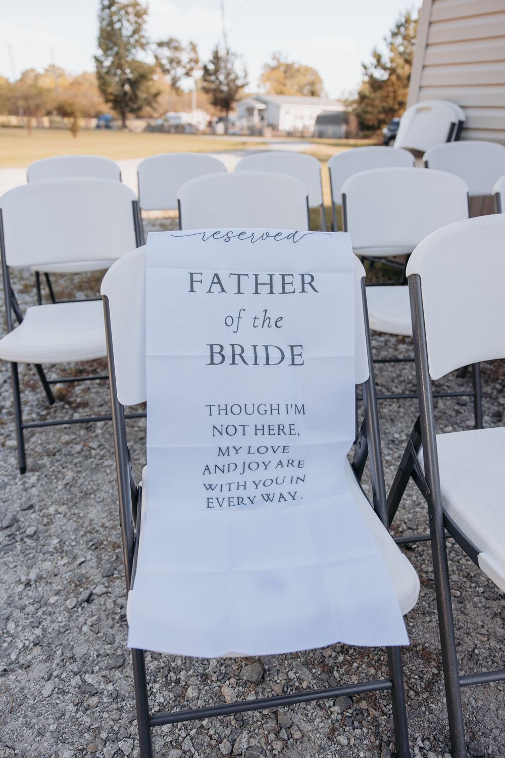 Empty white chairs at a wedding ceremony with signs reading 'Father of the Bride' and memorial messages.