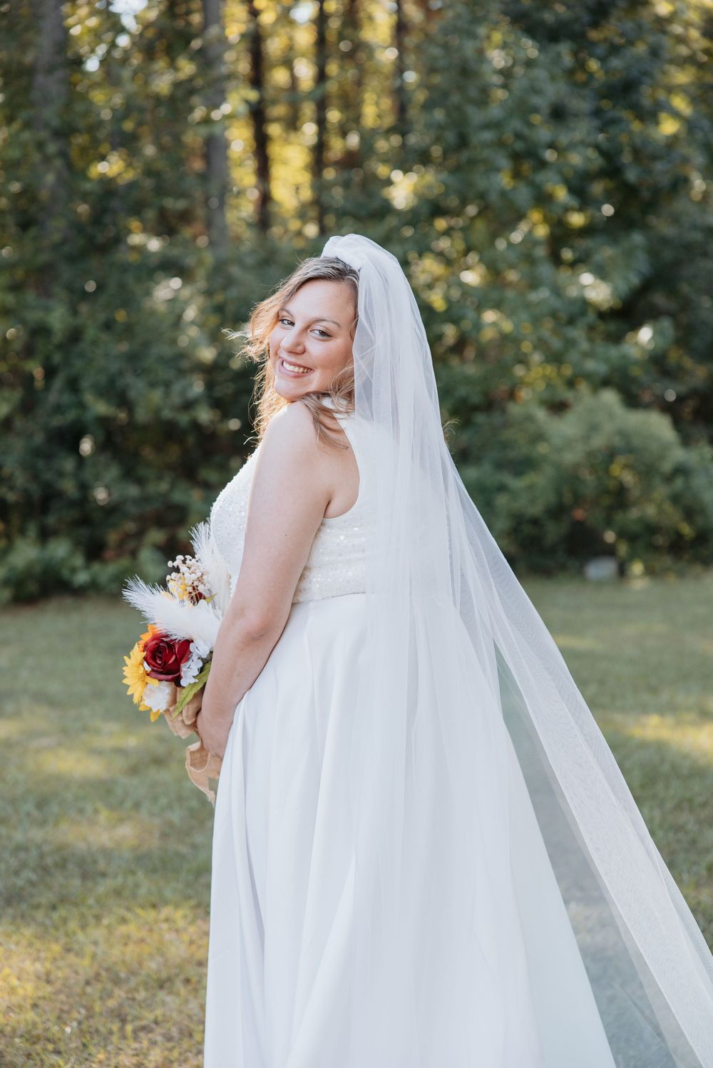 Bride in white wedding gown with flowing veil poses in outdoor setting.