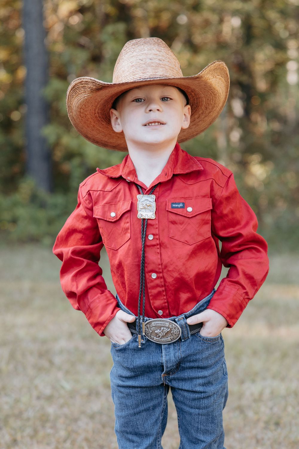 Young person in western attire featuring a red jacket, jeans, and cowboy hat posing outdoors.