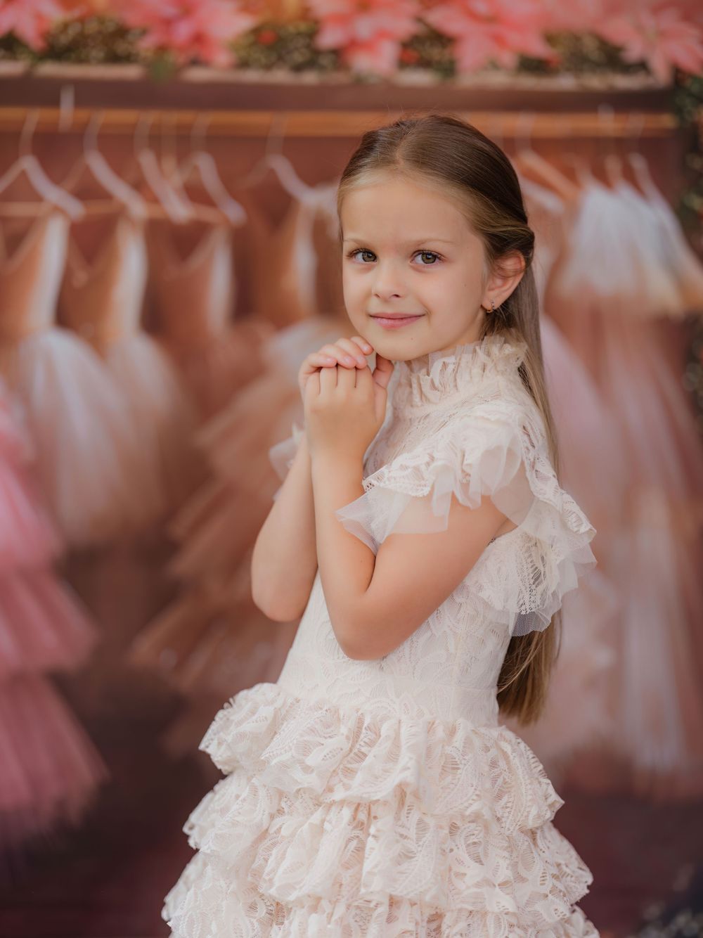 A child wearing a white lace dress with ruffles poses gracefully against a soft pink background.