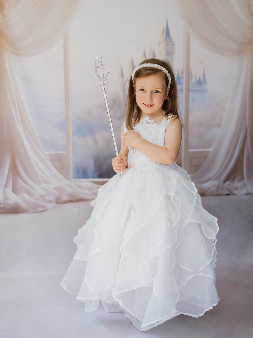 A child in a flowing white princess dress holds a sparkly wand against a dreamy castle backdrop with sheer curtains.