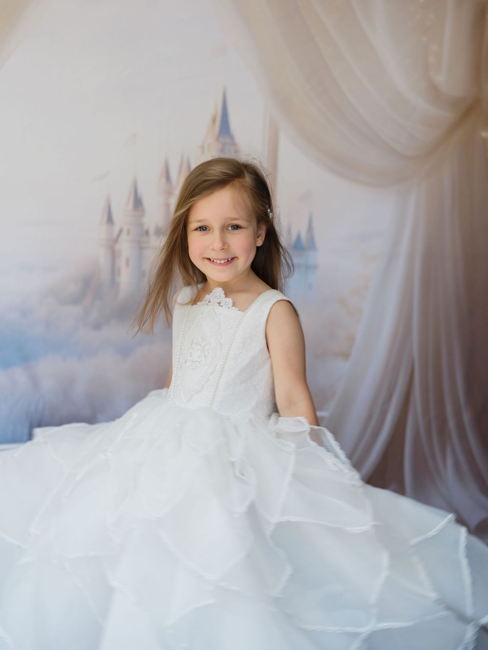 A young child wearing a flowing white dress poses gracefully against a dreamy backdrop with sheer curtains and soft lighting.