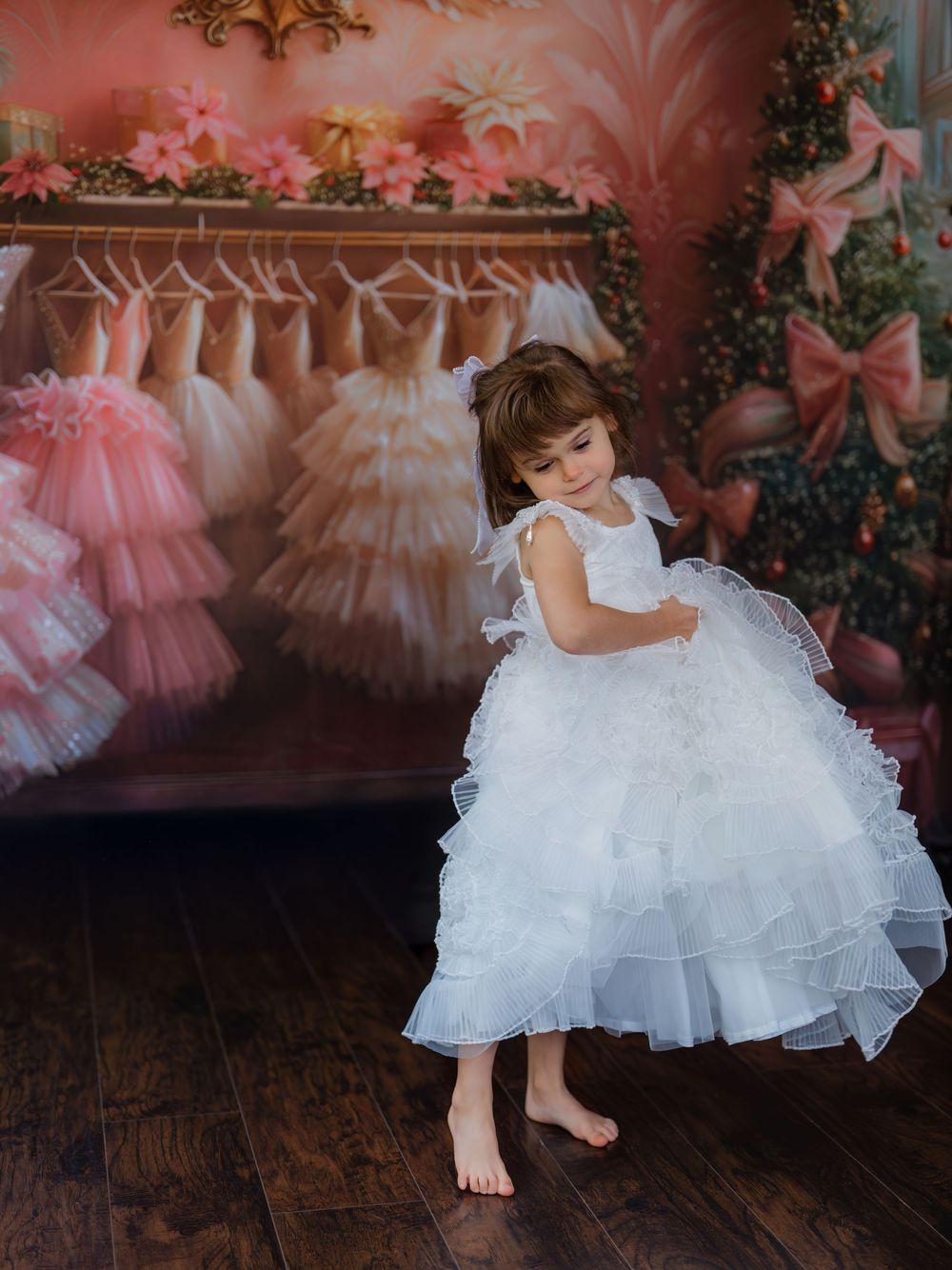 Young child in a fluffy white tulle dress twirls on a wooden floor against a pink backdrop with hanging dresses.