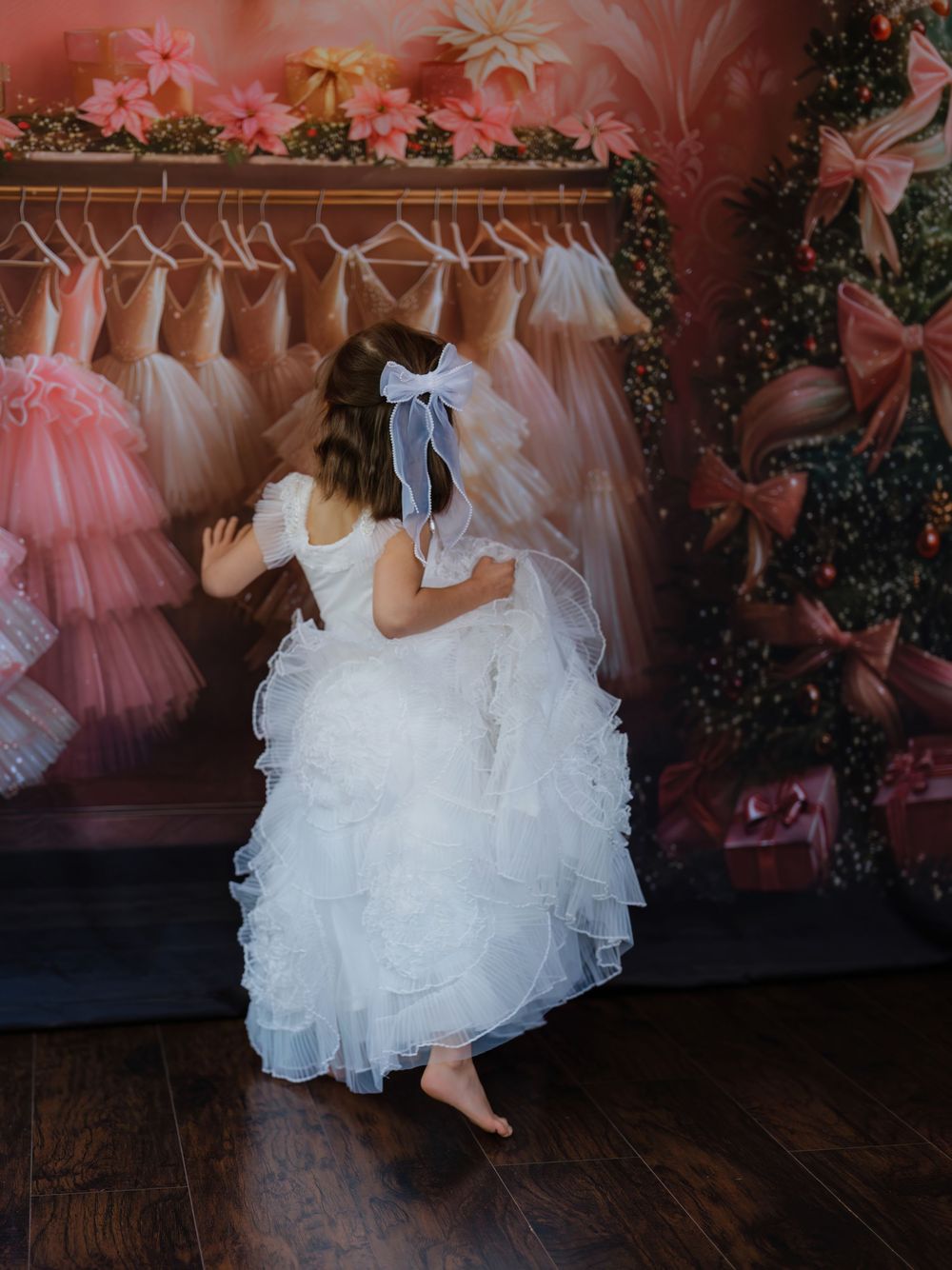 A wedding party wearing pink and white dresses poses together in front of a floral backdrop on dark hardwood floors.