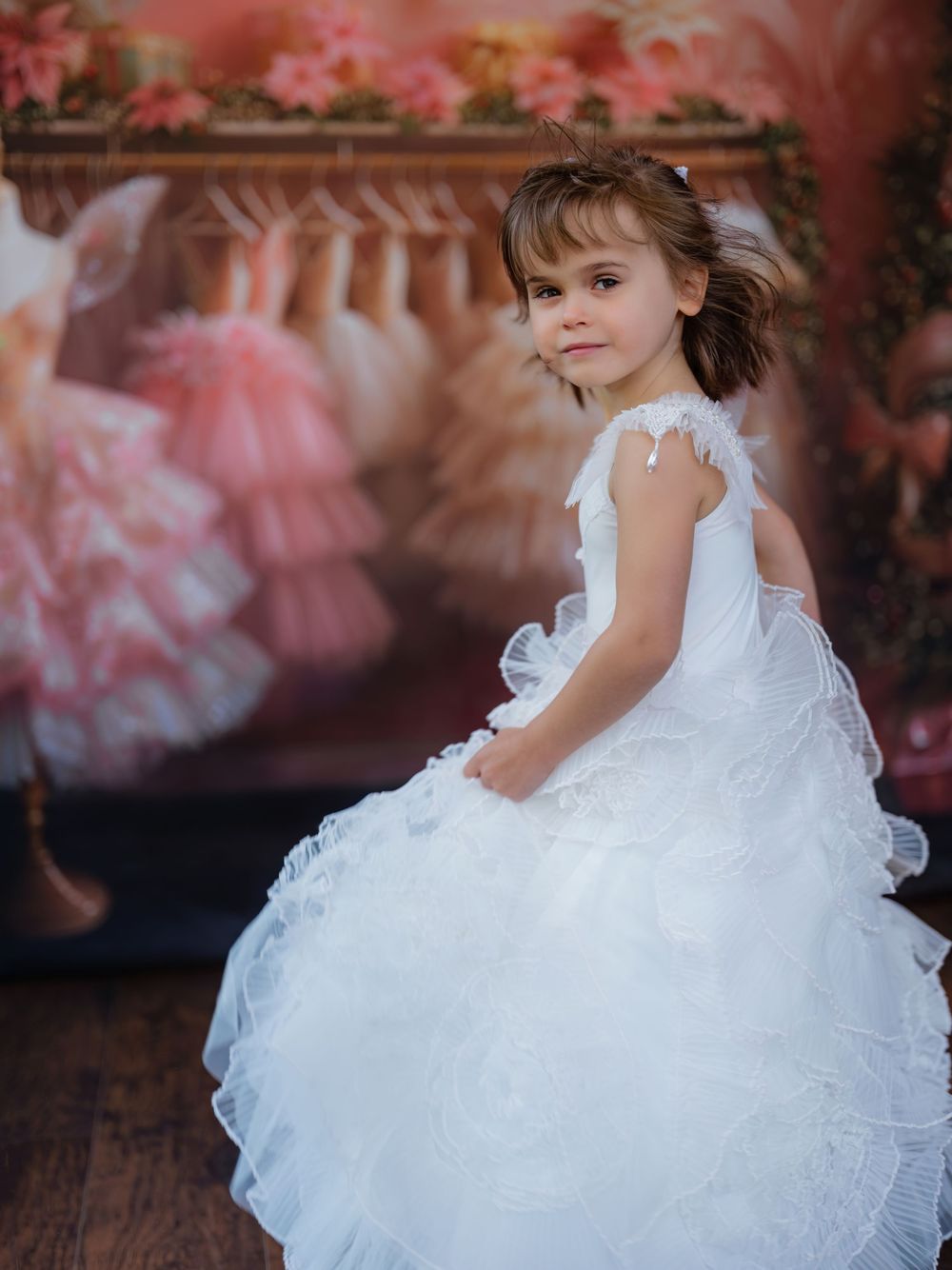 Child wearing white ruffled dress sits gracefully on dark wooden floor with pink tulle dresses in background.