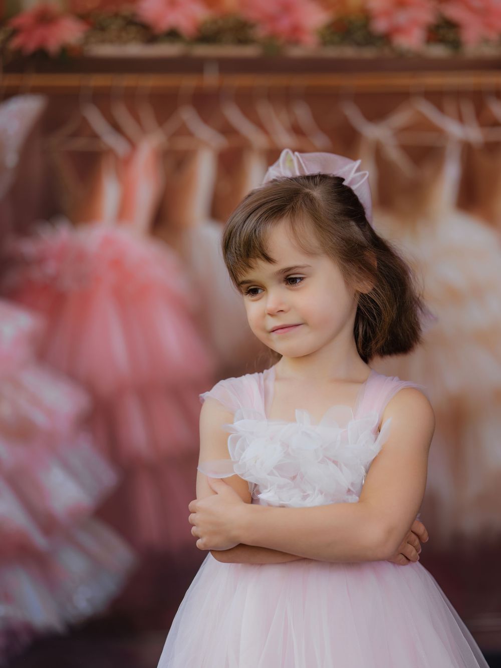 A child in a soft pink tulle dress with lace details poses against a dreamy pink floral backdrop.