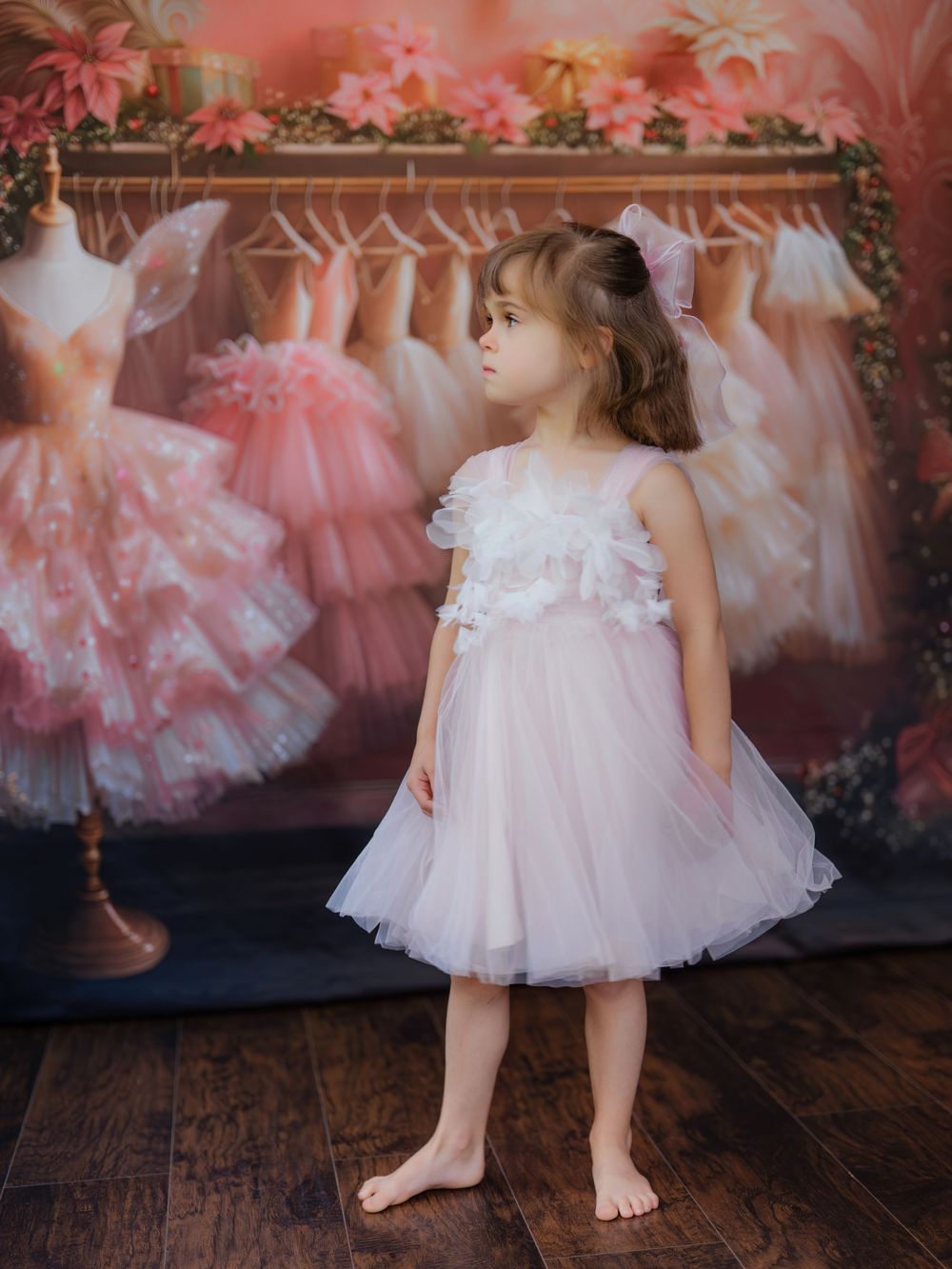 Small child in delicate white dress poses against pink background with tulle dresses displayed behind.