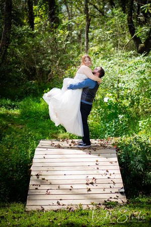 A couple dances romantically on a wooden bridge surrounded by lush green forest and fall leaves.