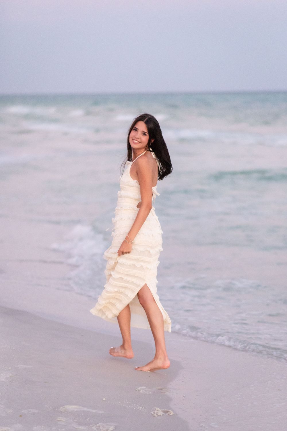 A person in a peach ruffled dress twirls joyfully on a serene beach with waves in the background.