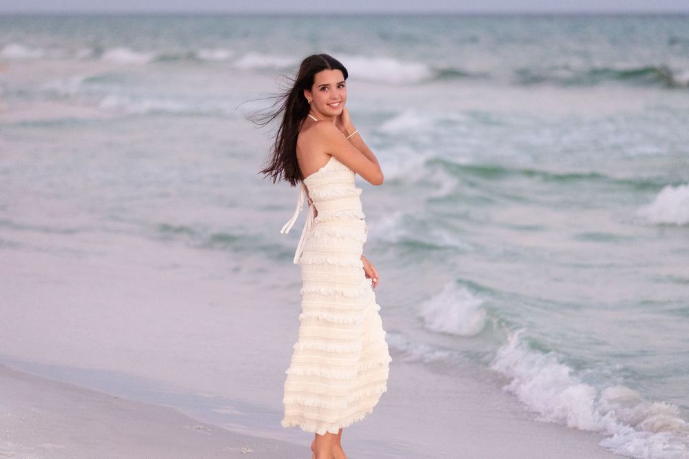 A woman in a flowing white dress walks along a beach at sunset creating a romantic silhouette against the ocean waves.
