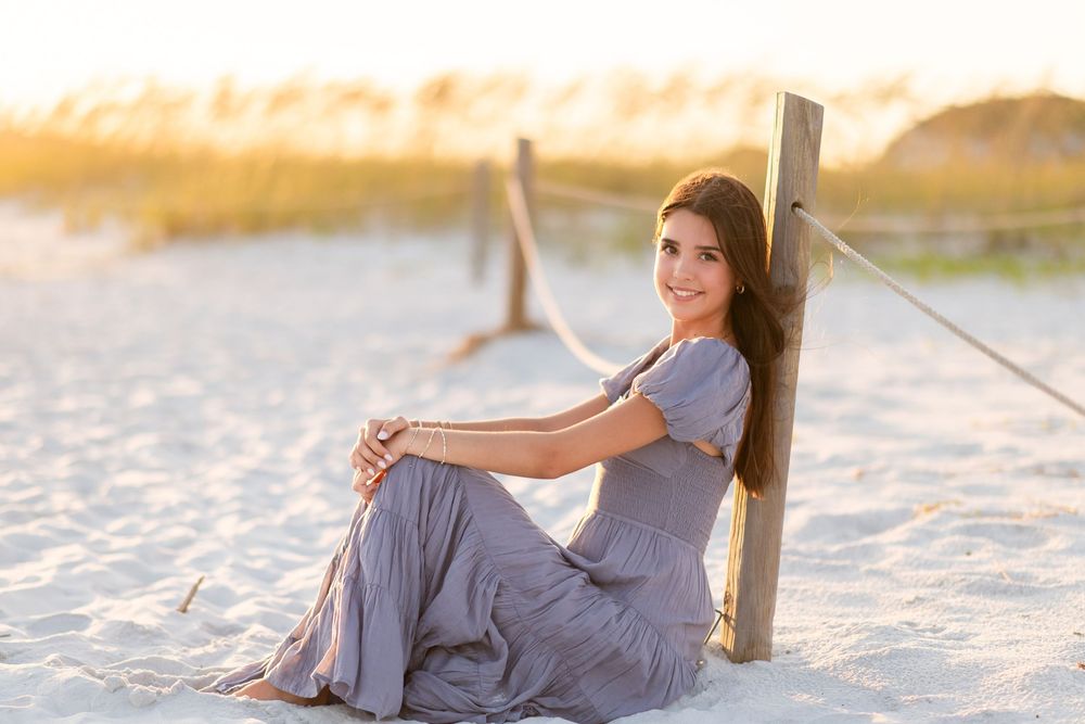 Woman in flowing gray dress poses gracefully on white sandy beach at sunset.