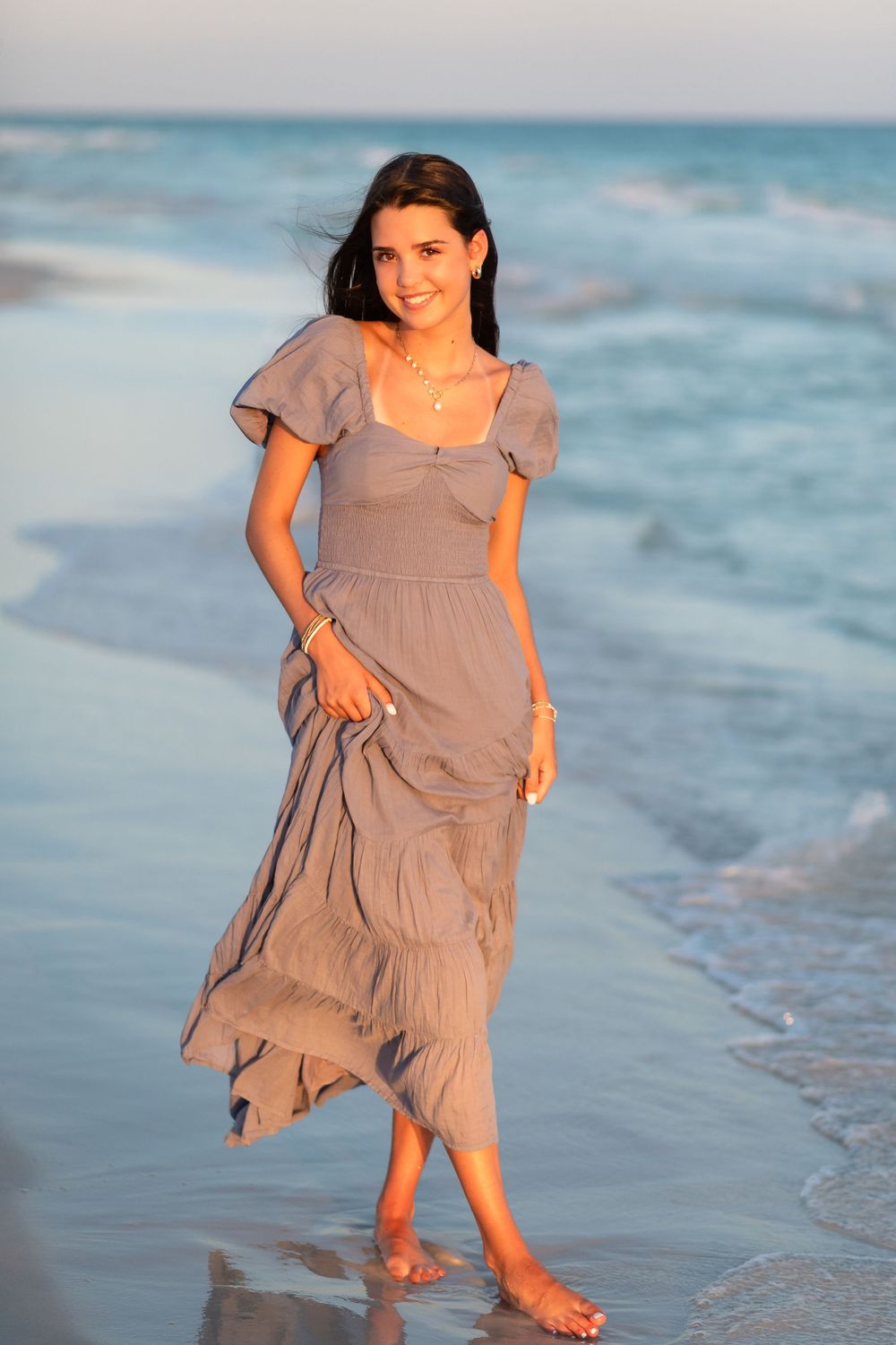 A young girl in a flowy taupe dress twirls and plays at the edge of the ocean waves during sunset.