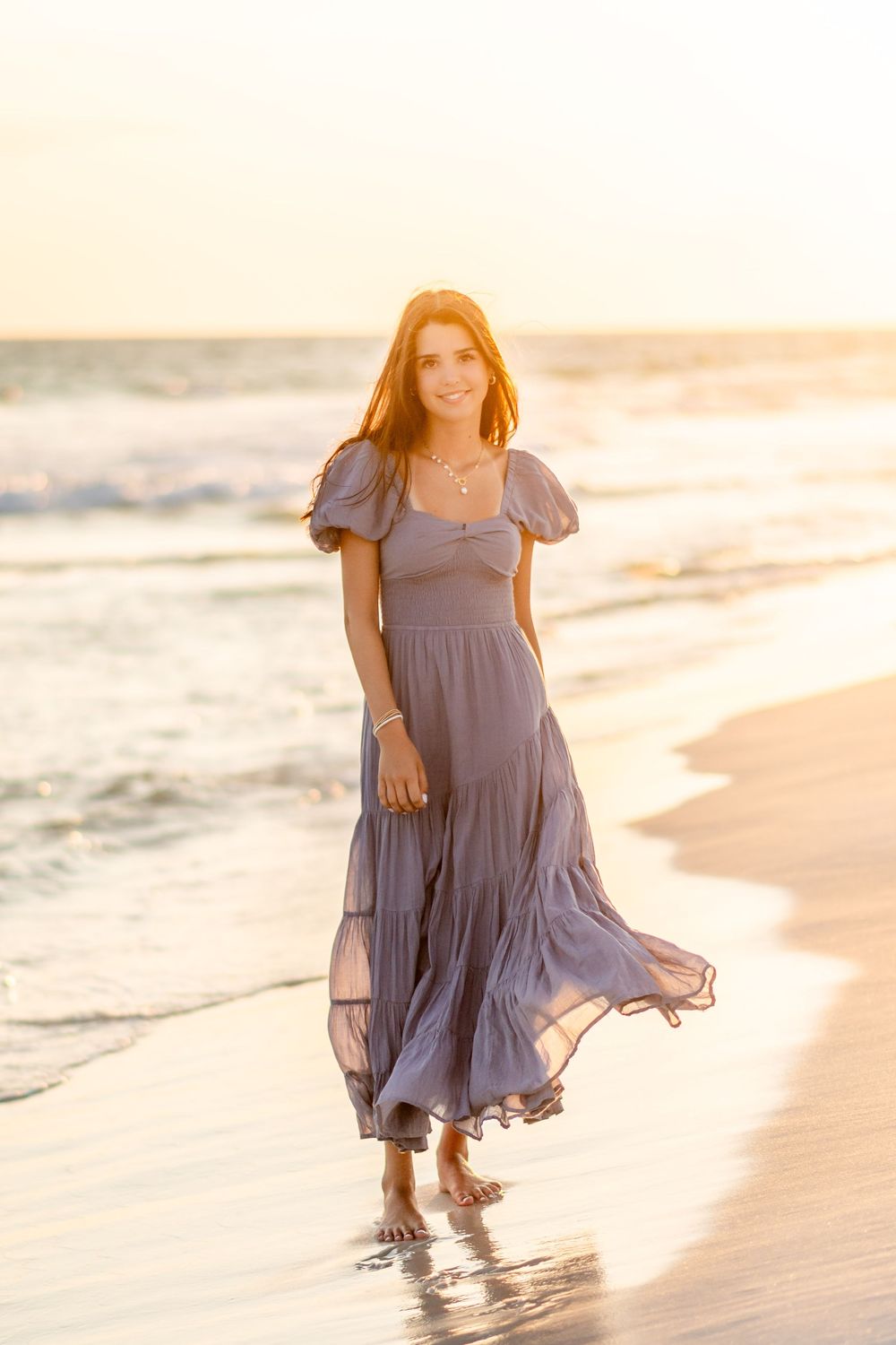 Child wearing a flowy lavender dress plays in the ocean waves at sunset on a sandy beach.