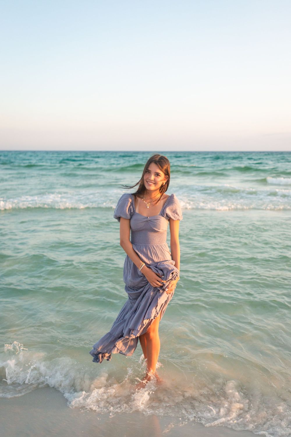 A person in a light blue dress splashes playfully in the turquoise ocean waves at sunset.