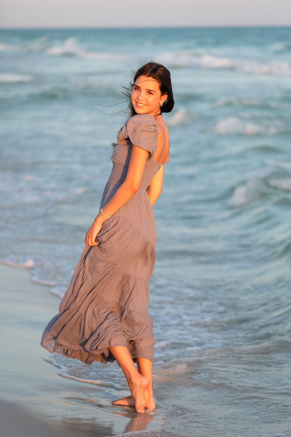 A person in a flowing taupe dress walks barefoot along the ocean shoreline as waves roll in during sunset.