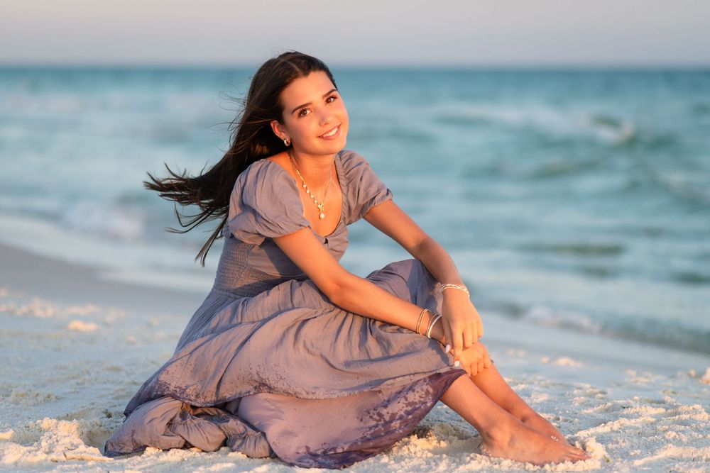 Woman in flowing mauve dress poses gracefully on sandy beach with ocean waves in background.