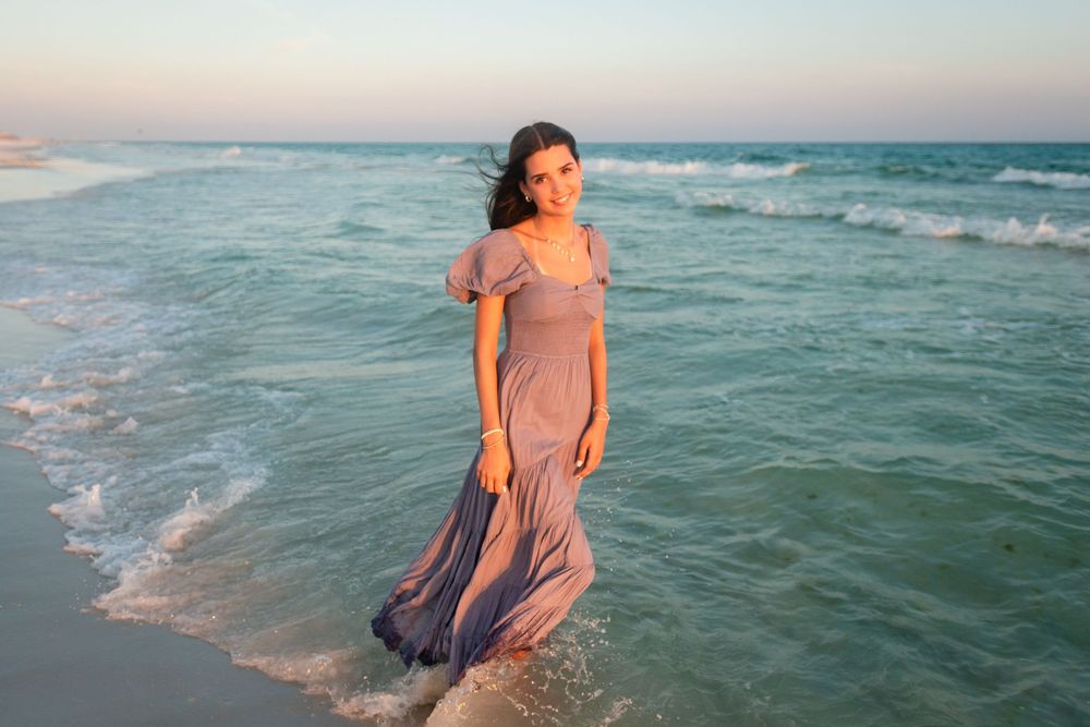 Someone in a flowing peach dress walks through turquoise ocean waves at sunset creating a dreamy coastal scene.
