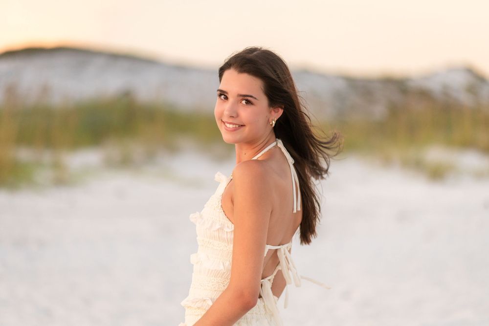 Woman in white dress smiling on beach with sunset lighting and ocean view.