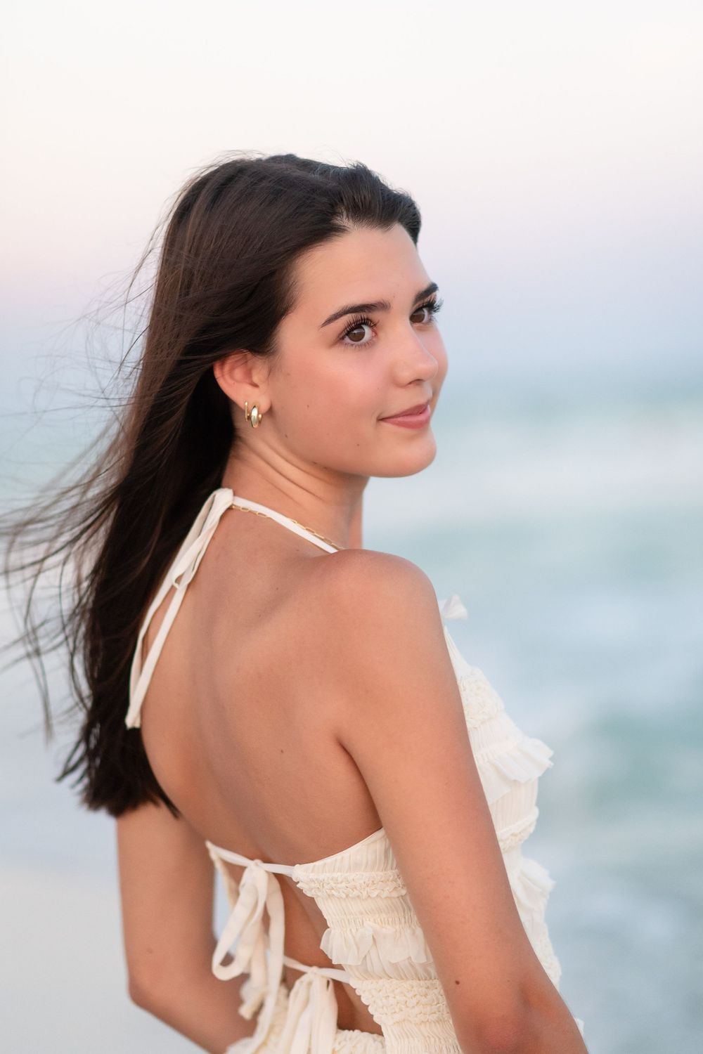 A woman in a white summer dress poses on a beach at sunset with the ocean in the background.