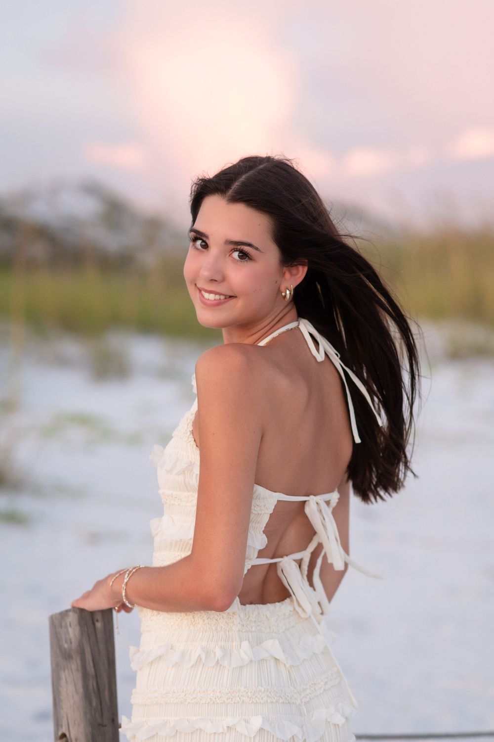 A woman in a white sundress poses on a beach at sunset with windswept hair and a joyful expression.