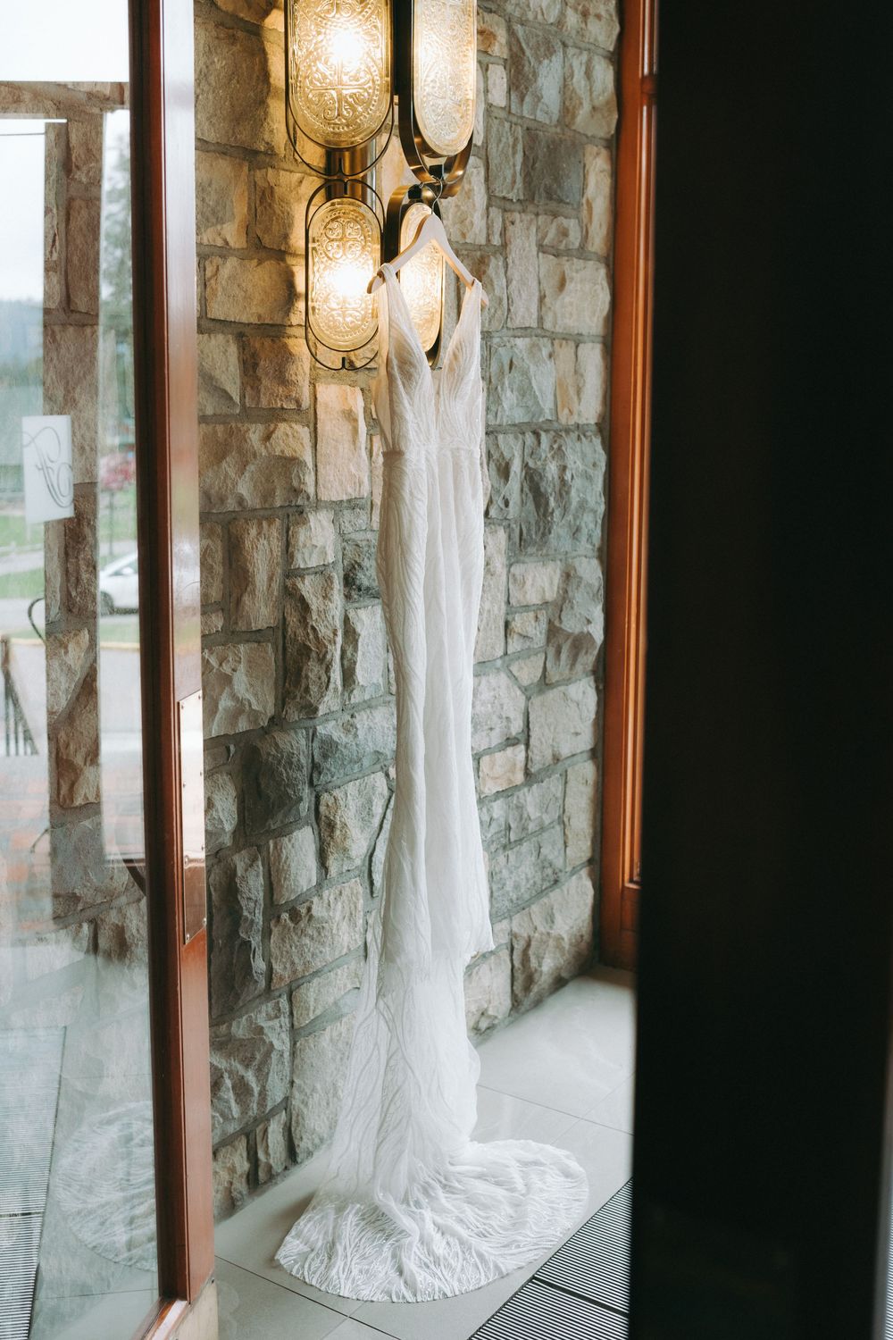 Elegant white wedding dress hanging against stone wall with warm lighting.