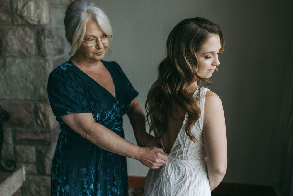 A woman in a teal sequin dress helps adjust a white wedding gown in a dimly lit room with stone walls.